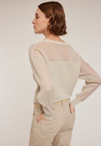 Beige ribbad tröja med genomskinliga randiga paneler vid axlar och ärmar, ihop med ljusbeiga byxor med bakfickor.