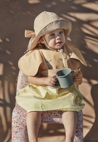 Bambino che indossa un cappello beige e un bavaglino abbinato con design a ciglia, tiene in mano una tazza grigia, seduto su una sedia con un motivo colorato, indossando una gonna a righe gialle.