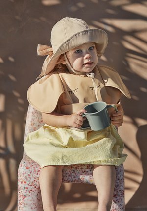 Barn iført en beige hat og matchende bib med øjenvippe-design, holder en grå kop, siddende på en farverig mønstret stol, iført en gul stribet nederdel.