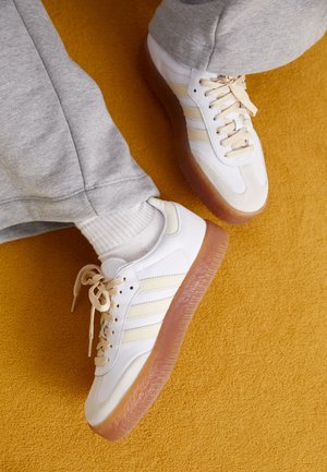 Witte leren sneakers met beige accenten, met een zool van gumrubber en drie tonale strepen, gecombineerd met grijze joggingbroeken.