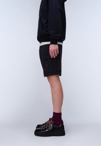 Inimene, kes kannab musti cargo-lühkse, musta jopet, burgundiapunaseid sokke ja musti matkastiilis jalanõusid, seisab profiilis ühevärvilise tausta ees.