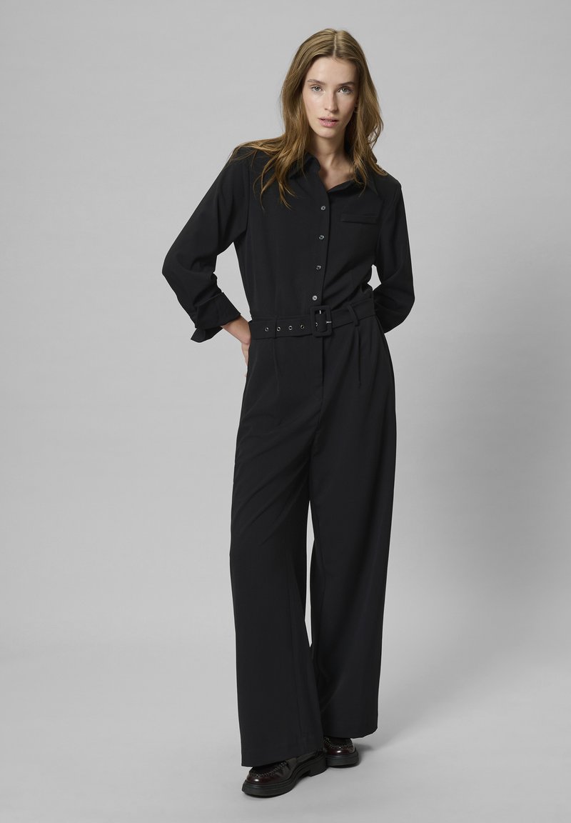 Fekete, hosszú ujjú overál gombos záródással, gallérral és széles lábakkal. Övvel ellátott derék és elülső zseb részletezéssel rendelkezik. Texturált anyag.