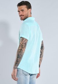 Hellblaues kurzärmliges Hemd mit relaxter Passform, das einen gefalteten Kragen und einen Rückensteg aufweist. Getragen mit hellblauen Jeans.