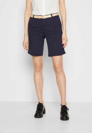 Kvinde iført mørkeblå knælange shorts med beige flettet bælte, hvid top og sorte snørestøvler, stående mod en ensfarvet baggrund.