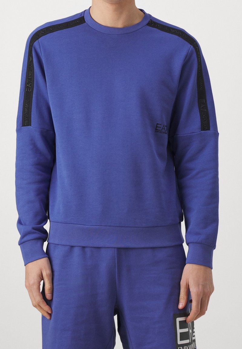 Uomo che indossa un felpa viola e pantaloni abbinati con strisce nere di marca sulle spalle e loghi sul petto e sulla coscia.