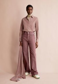 Camicia a righe con bottoni, jeans rosa a vita alta e sneakers con dettagli verdi. Tiene una soffice sciarpa rosa chiaro.