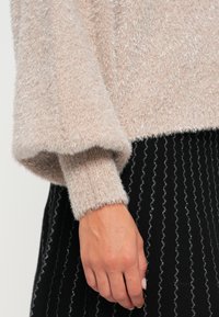 Pulover roz deschis dintr-un material moale, cu fire argintii, cu manșete cu textură, purtat peste o fustă neagră cu dungi argintii în formă de valuri. O mână este vizibilă pe mânecă.