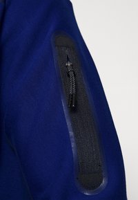 Maglione blu navy in tessuto strutturato, con tasca con zip sulla manica, accentuata da un elastico nero e un tirante per zip.