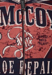 Sudadera con cremallera en rojo y azul marino con estampado gráfico, que presenta el texto "MCCOY", textura desgastada y cremallera plateada. Tejido suave.