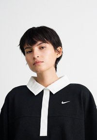 Mladý človek s krátkymi čiernymi vlasmi, oblečený v čiernej dlhoprstnej košeli s bielym golierom a logom Nike na hrudníku, na jednoduchom pozadí.