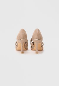 Chaussures à talons hauts en suède beige avec des talons fins et des lanières décoratives, présentées de dos sur un fond blanc uni.