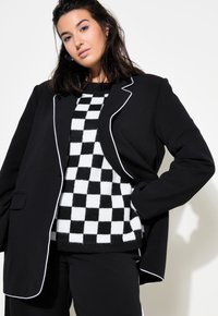 Černý blazer s bílým lemem přes černobíle kostkovaný svetr. Modelka ho kombinuje s černými kalhotami, čímž předvádí vrstvený vzhled.
