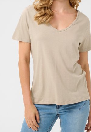 Vrouw met een beige T-shirt met korte mouwen en V-hals en een blauwe spijkerbroek, waarbij het bovenlichaam en een deel van de armen en taille zichtbaar zijn.