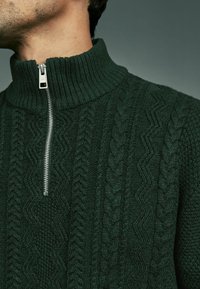Jersey de punto verde con cuello alto de canalé y cierre de media cremallera, con textura en relieve de trenzado y patrones en zigzag en toda la prenda.