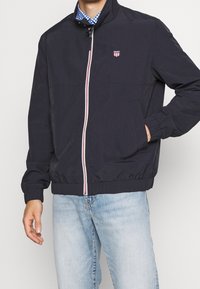 Granatowa kurtka typu windbreaker z czerwono-białym zamkiem błyskawicznym, elastycznymi mankietami i małym logo. Posiada boczne kieszenie oraz stójkę.