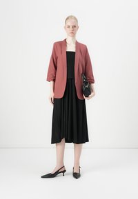 Sacou burgundy cu mânecile suflecate peste o rochie neagră fără mâneci. Pantofi cu toc slingback negri și o geantă mică neagră completează look-ul.