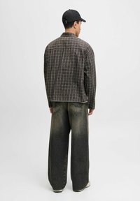 Een persoon staat met de rug naar de camera, gekleed in een geruite shirt en wijde vervaagde jeans, aangevuld met een zwarte pet tegen een effen achtergrond.