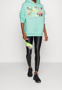 Felpa verde menta con stampa grafica, abbinata a leggings neri lucidi con un motivo astratto colorato e dettagli in neon verde. Sneakers nere.
