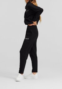 Woman in black matching tracksuit and white sneakers poses sideways, hands resting on lower back against plain gray background.