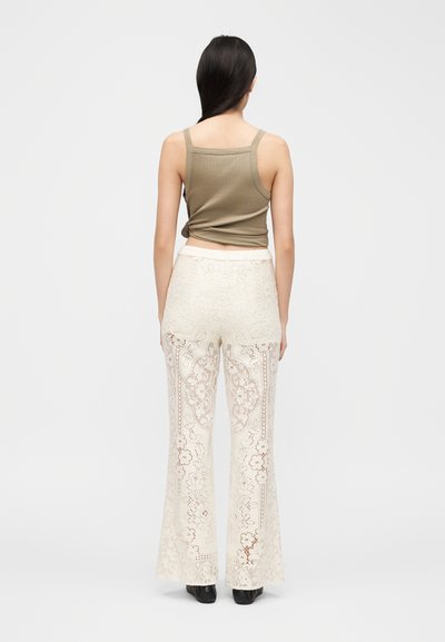 Γυναίκα με μακριά σκουρόχρωμα μαλλιά φοράει αμάνικη ελιάς μπλούζα και κρεμ δαντελωτό φαρδύ παντελόνι ψηλόμεσο, στέκεται με την πλάτη προς τα εμπρός σε απλό φόντο.