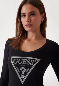 Musta pitkähihainen t-paita pyöreällä pääntiellä, jossa on hopeinen strassilogotriangeli keskellä.