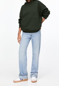 Felpa oversize verde scuro con tasca frontale e cappuccio regolabile, abbinata a jeans a gamba larga blu chiaro e sneakers bianche con strisce blu.