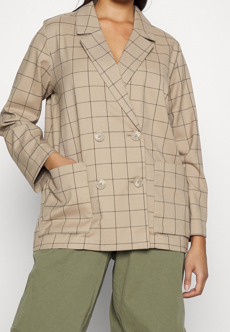 Femme portant un blazer beige croisé à carreaux noirs en motif quadrillé avec de grandes poches avant, associé à un pantalon vert olive.