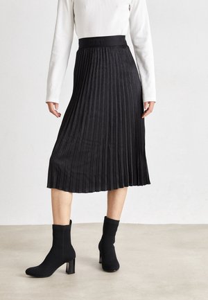 Femme portant une jupe midi plissée noire, un haut blanc à manches longues et des bottines noires à talons, debout sur un sol gris clair.