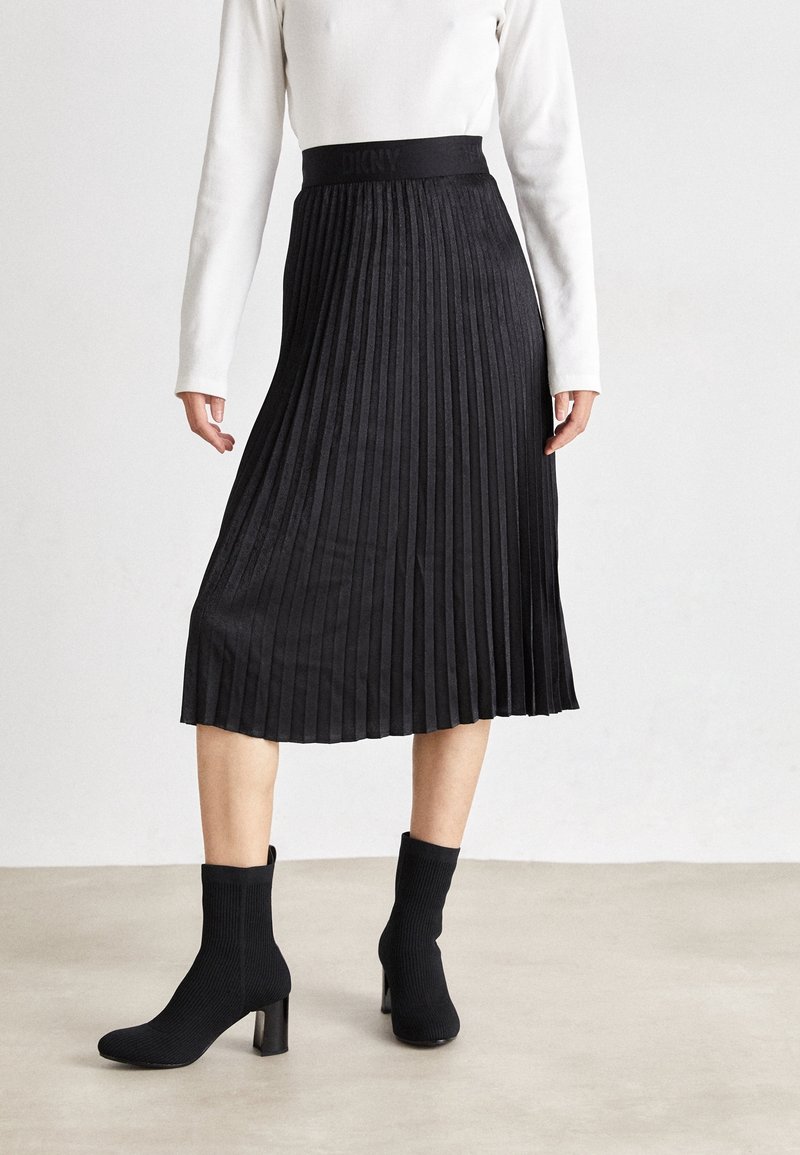 Femme portant une jupe midi plissée noire, un haut blanc à manches longues et des bottines noires à talons, debout sur un sol gris clair.