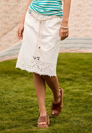 Žena v bielej vyšívanej midi sukni s pleteným opaskom, tyrkysovom pruhovanom topu a hnedých platformových sandáloch kráča po tráve.