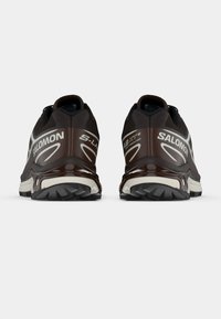 Brūni sporta apavi ar melniem akcentiem, glīts dizains, viegla materiāla, teksturētas zoles un izteikta "SALOMON" zīmola identifikācija.
