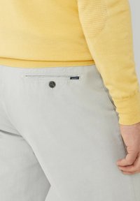 Pantaloni grigi chiari realizzati in tessuto morbido, con una tasca sul retro dotata di bottone e un piccolo etichetta del marchio. Abbinati a un maglione giallo.