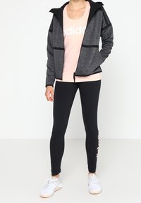 Chaqueta gris con cremallera y acentos negros, camiseta rosa con un logo, leggings negros con letras rosas y zapatillas deportivas blancas.