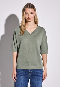 T-shirt z dekoltem w serek i krótkim rękawem w oliwkowym kolorze, wykonany z miękkiego materiału, zestawiony z niebieskimi dżinsami. Prosty, luźny krój.