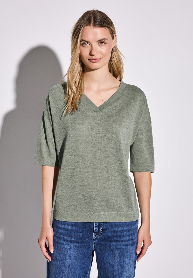 T-shirt z dekoltem w serek i krótkim rękawem w oliwkowym kolorze, wykonany z miękkiego materiału, zestawiony z niebieskimi dżinsami. Prosty, luźny krój.