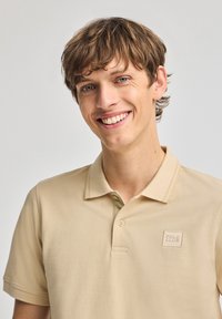 Ljusa beige polo-shirt med en texturerad tyg, korta Ärmar och en sydd logopatch på vänster bröst. Knappslå vid halsen.
