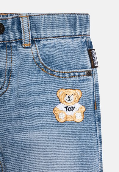 Svetlomodré džínsové šortky s vyšitou nášivkou medvedíka a textom "TOY". Doplnky zahŕňajú žlté prešitie a čiernu logo nálepku.