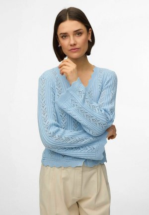 Vrouw met kort bruin haar die een lichtblauwe gebreide trui met een geschulpte rand en beige hoge taillebroek draagt, poserend met gevouwen armen.