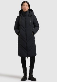 Lang sort pufferjakke med chevron-syninger, en høj krave og justerbare snore. Båret over sorte leggings og ankelstøvler.