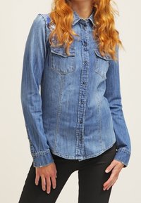 Vrouw in een getailleerde blauwe spijkerblouse met borstzakken en zwarte broek, staand met de handen ontspannen aan de zijkanten.