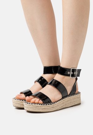 Sandales en cuir verni noir avec deux larges sangles et une sangle au niveau de la cheville, dotées d'une semelle en jute tressé et d'un détail de boucle métallique.