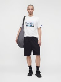 Homme portant un t-shirt blanc avec un imprimé camion de glace, short noir, baskets noires, chaussettes noires et un sac bandoulière gris, debout sur fond blanc.