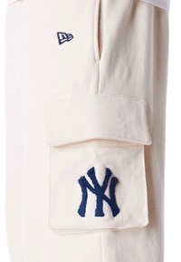 Beige cargo bukser lavet af blødt stof, med et marineblåt broderet logo og en oversized lomme på låret. Minimalistisk design.