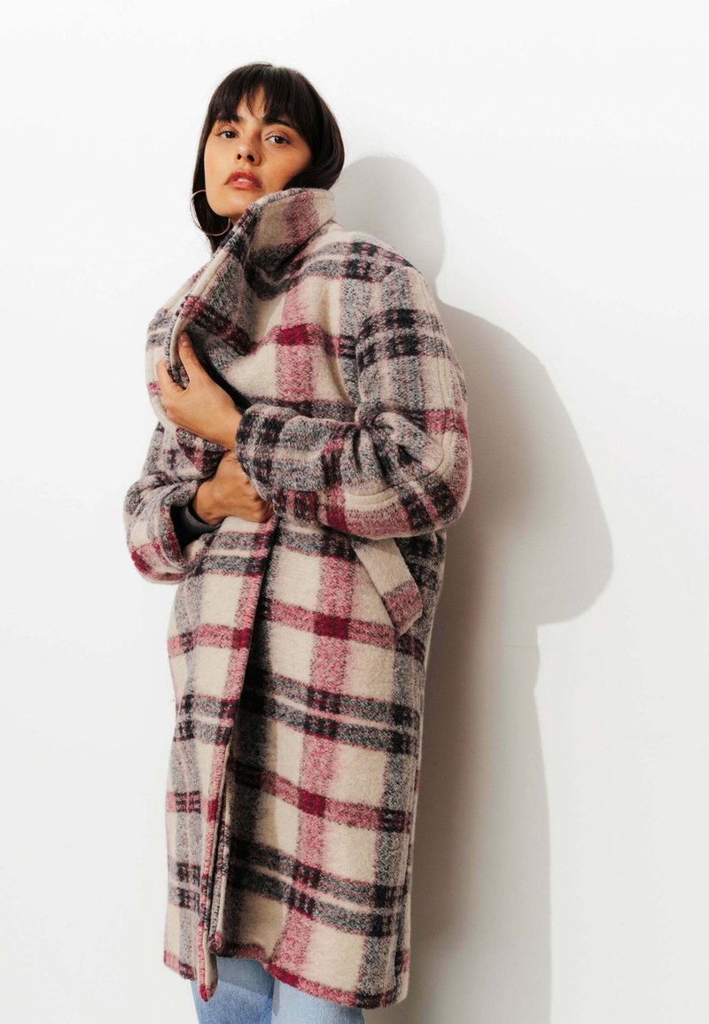 Manteau en mélange de laine à carreaux avec un col haut, dans des teintes de crème, rouge et gris. Coupe oversize, avec des manches amples et des poches latérales.