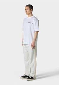 Hvit oversized t-skjorte med svart logo på brystet, paret med hvite bukser med sidelister, og svarte sneakers med beige snører.
