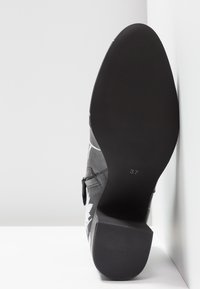 Zapato de cuero negro con bordado blanco, tacón alto, suela plana y tamaño marcado como 37 en la parte inferior. Diseñado con puntera afilada.
