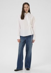White collared shirt with a side tie detail, featuring long sleeves and a relaxed fit, paired with wide-leg blue denim jeans.