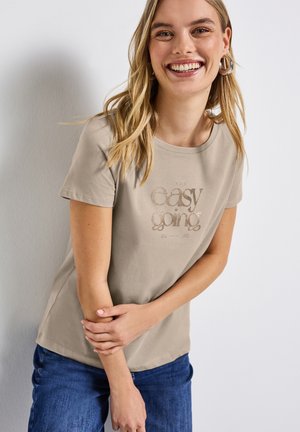 T-shirt en coton beige à manches courtes, col rond, avec un texte métallique qui dit "easy going". Associé à un jean en denim bleu.