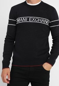Pull noir en maille avec des rayures horizontales blanches et le logo "ARMANI EXCHANGE" ; encolure arrondie ; poignets et ourlet côtelés avec un accent rouge.