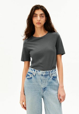 Frau mit langen dunklen Haaren, die ein dunkelgraues T-Shirt mit kurzen Ärmeln trägt, das in hellblaue hoch taillierte Jeans gesteckt ist, stehend vor weißem Hintergrund.